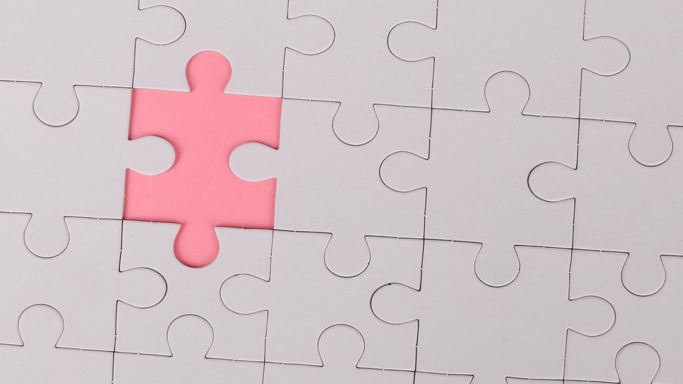 Close up of white jigsaw puzzle in assembled state with one piece missing revealing pink background
